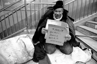 老人一边在寒风中乞讨，一边拿起一个失物招领牌向过路人展示。
